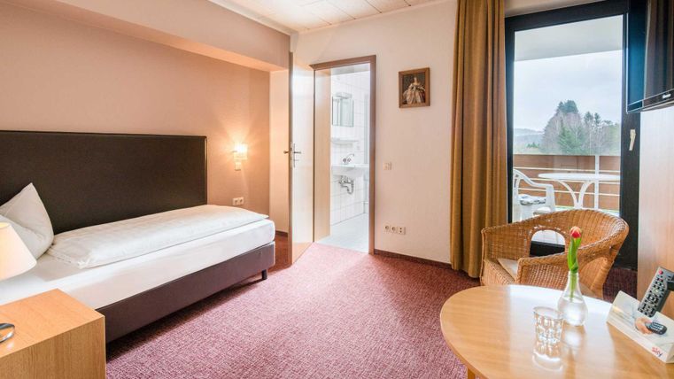 Ein helles Hotelzimmer mit einem Einzelbett und einem gemütlichen Sessel. Es gibt einen kleinen Tisch und Zugang zu einem Balkon.