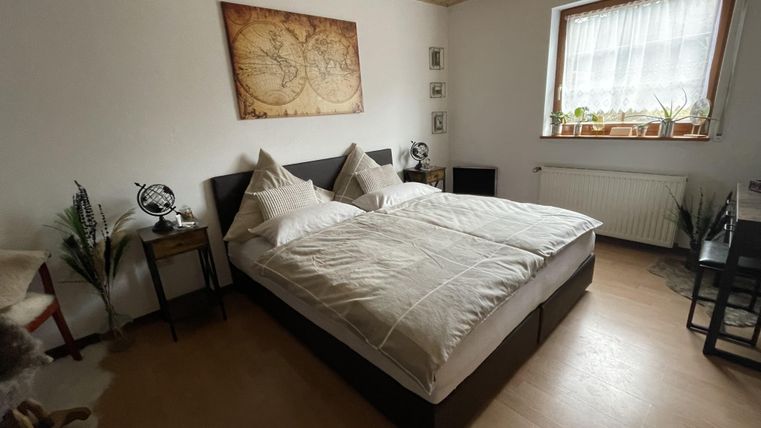 Ein gemütliches Schlafzimmer mit einem großen Bett und einer warmen, einladenden Atmosphäre. An den Wänden hängen dekorative Bilder, und das Licht kommt durch ein Fenster mit Vorhängen.