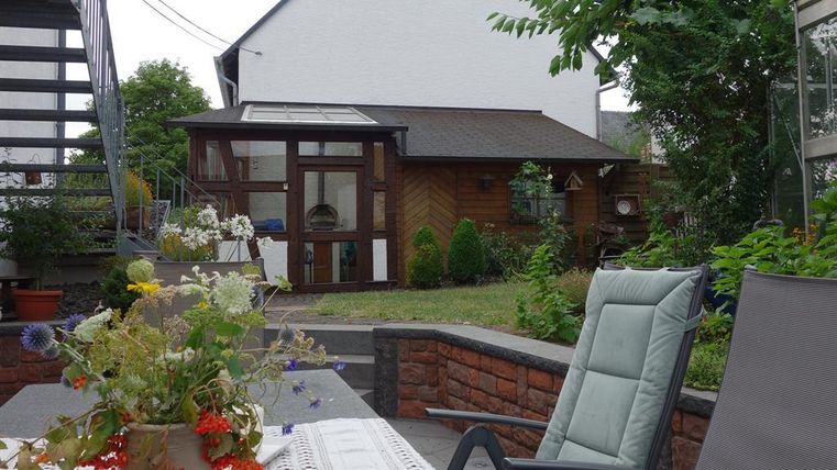 Ein gemütlicher Garten mit einem Tisch und Stühlen. Im Hintergrund sieht man ein Holzgebäude und üppige Pflanzen.