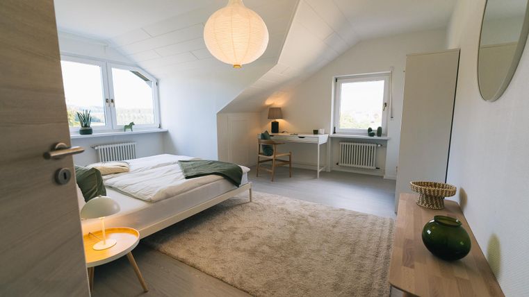 Ein schlafzimmer mit einem modernen Design und gemütlicher Atmosphäre. Es gibt ein Bett, einen Schreibtisch und große Fenster mit Ausblick.