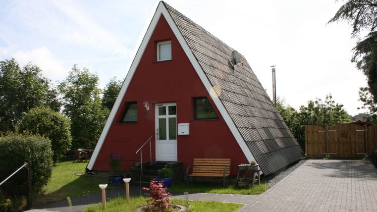 Ein rotes, dreieckiges Ferienhaus mit einem schrägen Dach. Umgeben von grünem Garten und einer Terrasse.