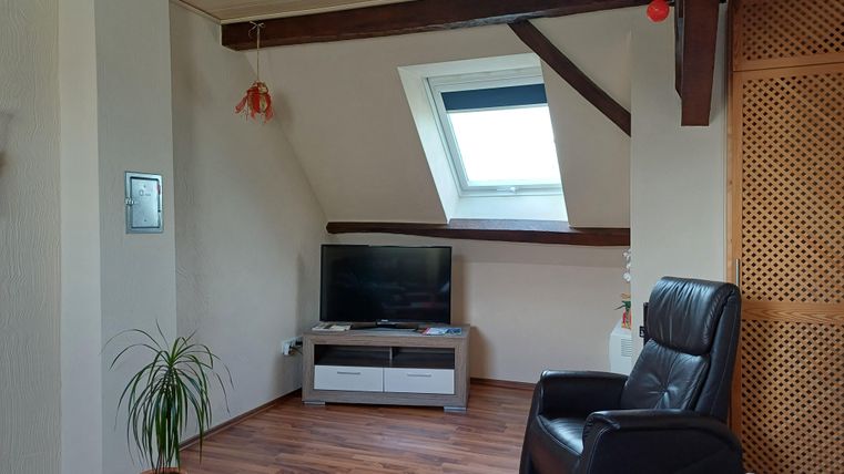 Ein gemütliches Wohnzimmer mit einem Fernseher und einem bequemen Sessel. Ein großes Dachfenster lässt viel Licht hereinfällen.