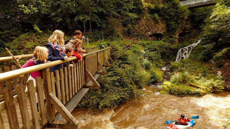 Eine Gruppe von Kindern und Erwachsenen steht auf einer Holzbrücke und schaut einem Kajakfahrer im reißenden Wasser zu. Im Hintergrund sind Bäume und ein kleiner Wasserfall sichtbar.