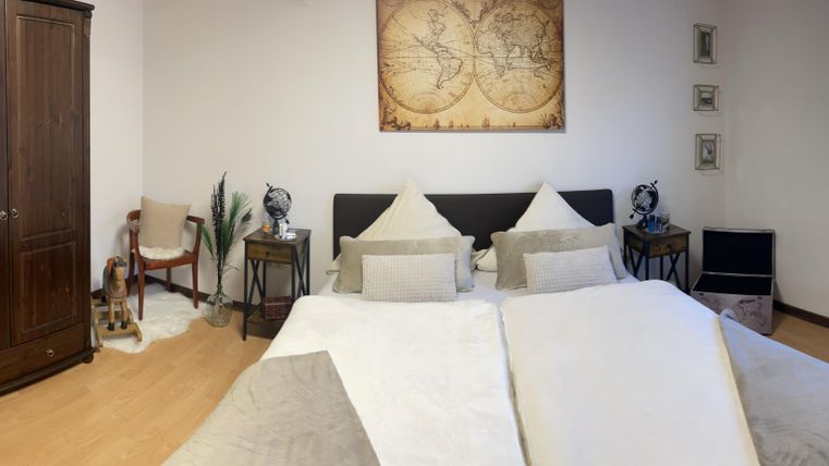 Ein gemütliches Schlafzimmer mit zwei Betten, einem großen Schrank und einem Fenster. Die Wände sind schlicht, und es gibt einige Pflanzen sowie dekorative Elemente im Raum.