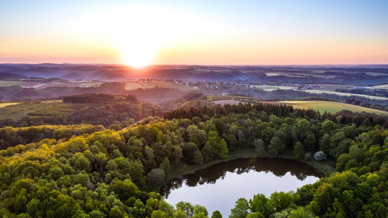 Luftaufnahme des Windsborn-Kraters mit See und Sonnenaufgang im Hintergrund.