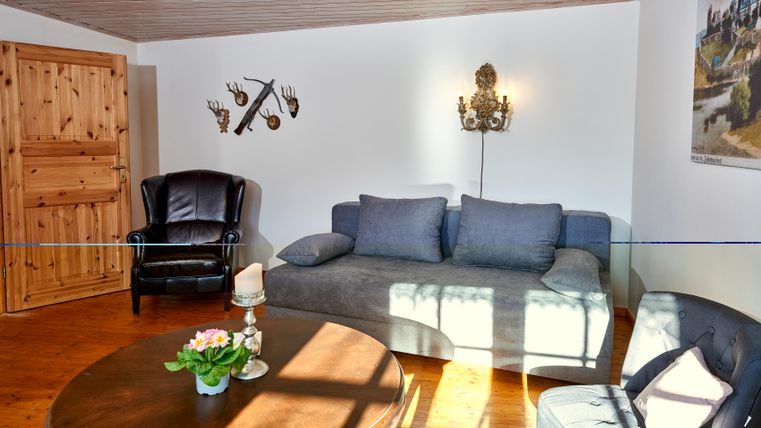 Ein gemütliches Wohnzimmer mit einem blauen Sofa und einem schwarzen Sessel. An der Wand hängen einige dekorative Elemente und das Licht fällt sanft auf den Tisch.