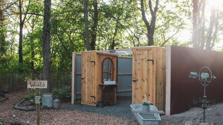 Ein rustikales, freistehendes Badezimmer im Wald mit Holzverkleidung und einem Spiegel. Es gibt eine rustikale Waschtisch-Installation und einen Weg aus Holzspänen.