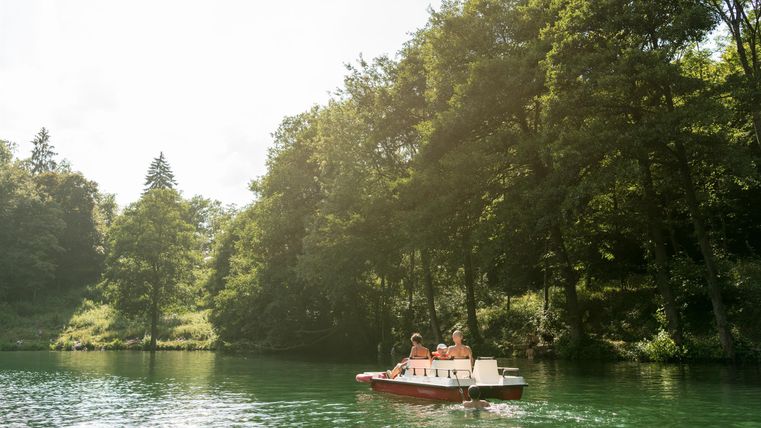 Ein Boot fährt auf einem ruhigen, grünen Gewässer. Umgeben von üppigen Bäumen und hellem Licht wirkt die Szene friedlich.