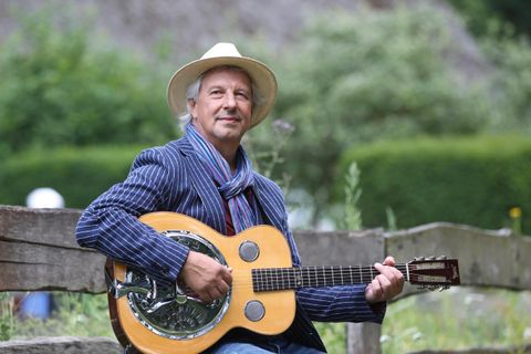Ein Mann sitzt auf einer Bank und spielt Gitarre. Er trägt einen Hut und hat ein freundliches Lächeln.