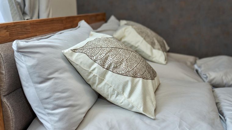 Ein gemütliches Bett mit hellen Kissen und einem schlichtem Holzrahmen. Die Kissen haben ein schönes Blattmuster und laden zum Entspannen ein.