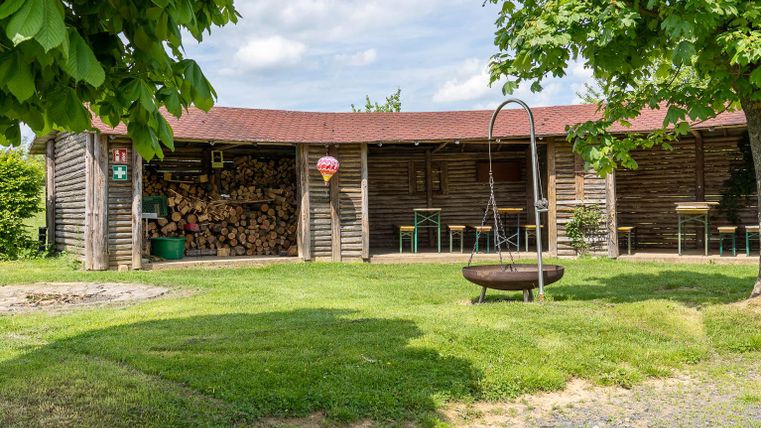 Eine gemütliche Holzhütte mit gestapeltem Brennholz. Der Garten ist grün und einladend, mit einem Schwenkstuhl und einem Feuerkorb.