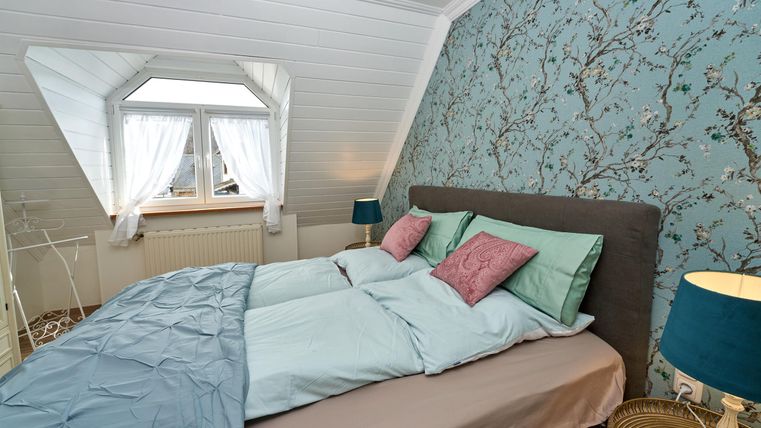 Ein gemütliches Schlafzimmer mit einem großen Bett und frischen Bettwäsche in sanften Farben. An den Wänden ist ein schöner Blumen-Tapeten-Muster zu sehen, und das Fenster lässt viel Licht herein.