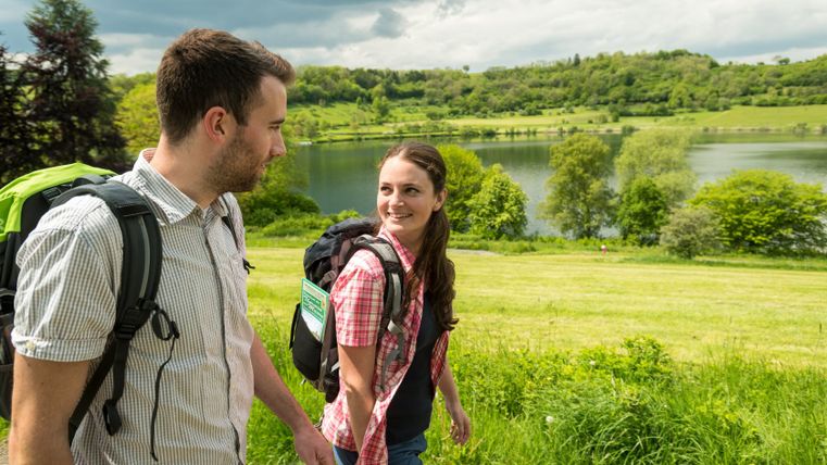 Zwei Wanderer am Schalkenmehrener Maar mit Rucksäcken, im Hintergrund der See und grüne Hügel.