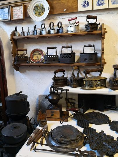 Eine Sammlung von antiken Eisenwaren und Küchengeräten steht auf einem Tisch und Regal. Die Objekte zeigen verschiedene Modelle von Bügeleisen und Kochgeschirr.