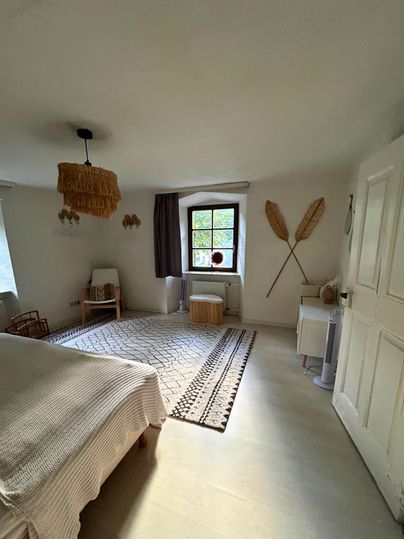 Ein helles Schlafzimmer mit einem großen Teppich und dekorativen Elementen an den Wänden. Die Einrichtung ist schlicht und modern, mit einem Fenster, das Tageslicht hereinkommen lässt.