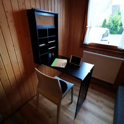 Ein moderner Schreibtisch mit einer weißen Stuhl in einem Holzraum. Auf dem Tisch liegen Schreibutensilien und ein Tablet, während das Licht durch ein Fenster strömt.