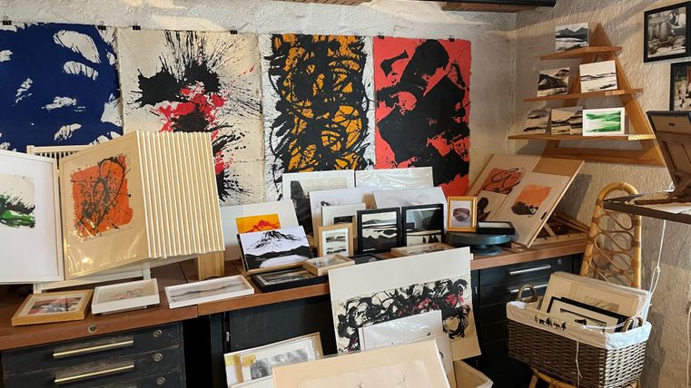 Ein kreatives Atelier mit verschiedenen Kunstwerken und Zeichnungen. Der Tisch ist voll mit Papieren und Kunstmaterialien, während bunte Bilder an der Wand hängen.