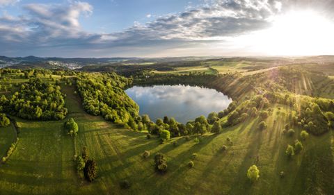 Eine malerische Landschaft mit sanften Hügeln und einem ruhigen klaren See. Die Sonne scheint durch die Wolken und beleuchtet die grünen Wiesen und Bäume.