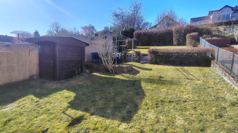 Ein sonniger Garten mit kurzem Gras und einem Holzschuppen. Im Hintergrund sind Hecken und einige Bäume zu sehen.