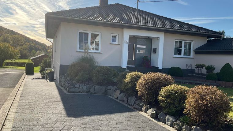 Ein gepflegtes Einfamilienhaus mit einem schönen Garten und einer gepflasterten Einfahrt. Die Umgebung ist von grünen Hügeln umgeben.