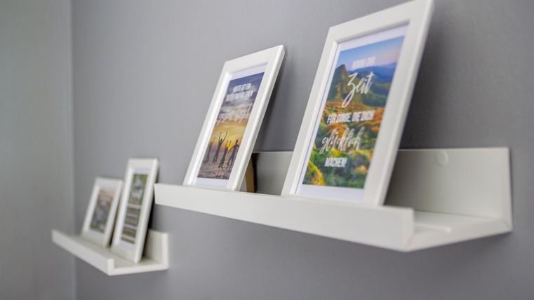 Rahmen mit Bildern stehen auf schlichten, weißen Regalen an einer grauen Wand. Die Bilder zeigen verschiedene Motive und verleihen dem Raum eine persönliche Note.