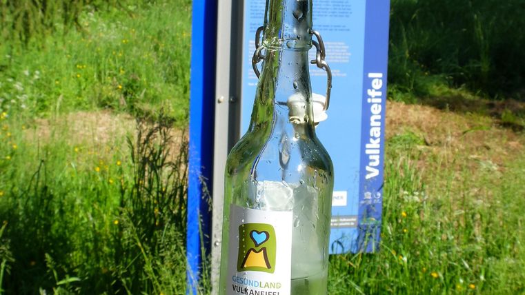 Eine Glasflasche mit einem Etikett steht im Vordergrund, während ein Informationsschild im Hintergrund sichtbar ist. Die Umgebung besteht aus grüner Wiese und Bäumen.