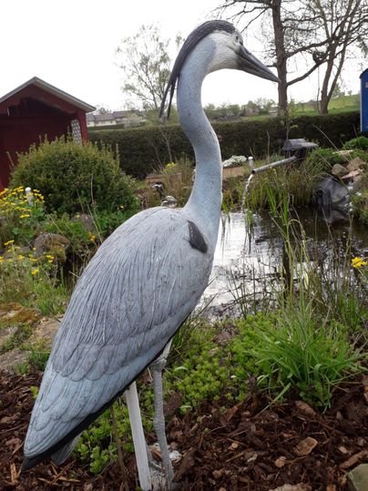 Eine fiktive Statue eines Graureihers steht neben einem Teich. Umgeben von Pflanzen und Blumen, verleiht sie dem Garten eine natürliche Atmosphäre.