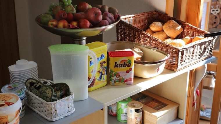 Ein Tisch mit einer Obstschale voller Äpfel und Trauben, und einem Korb mit Brötchen. Daneben steht eine Auswahl an Getränken und Lebensmitteln.