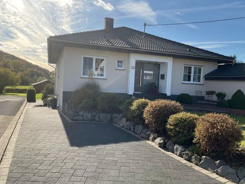 Ein gepflegtes Einfamilienhaus mit einem schönen Garten und einer gepflasterten Einfahrt. Die Umgebung ist von grünen Hügeln umgeben.