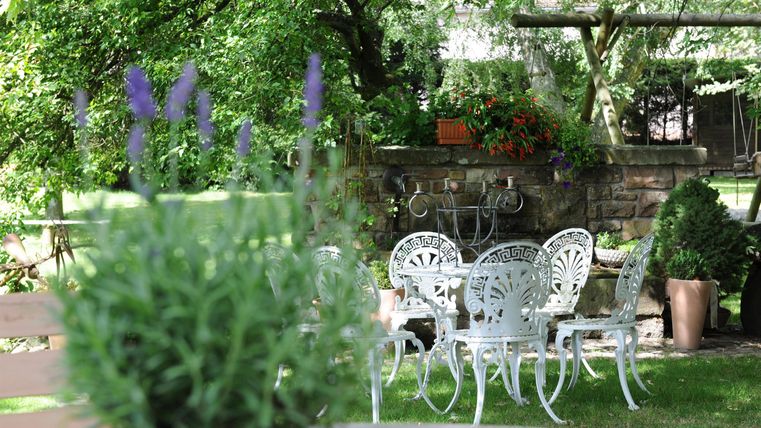 Ein einladender Garten mit weißen Gartenmöbeln und bunten Pflanzen. Im Vordergrund blühen Lavendelblüten.