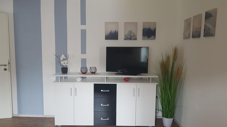 Ein modernes Wohnzimmer mit einem weißen TV-Schrank und einem Flachbildfernseher. An der Wand hängen dekorative Bilder und eine grüne Pflanze steht in der Ecke.