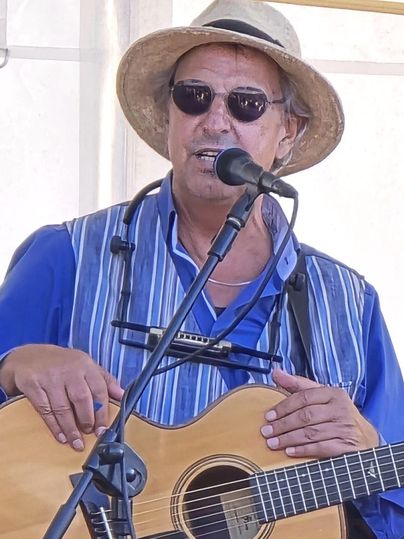 Ein Mann mit Sonnenbrille und Hut spielt auf einer Akustikgitarre. Er steht vor einem Mikrofon und singt, während er in einem lässigen Hemd gekleidet ist.
