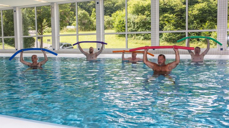 Eine Gruppe von Menschen macht Aquafitness im Becken. Sie halten bunte Schwimmnudeln und üben gemeinsam.