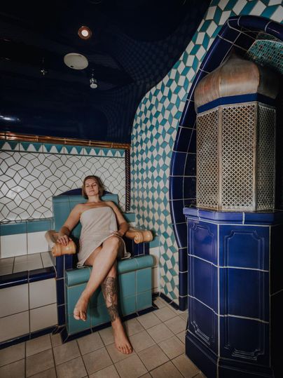 Eine Person sitzt in einem blauen Wellnessbereich auf einem bequemen Stuhl. Die Wände sind mit bunten Fliesen dekoriert, die eine entspannende Atmosphäre schaffen.