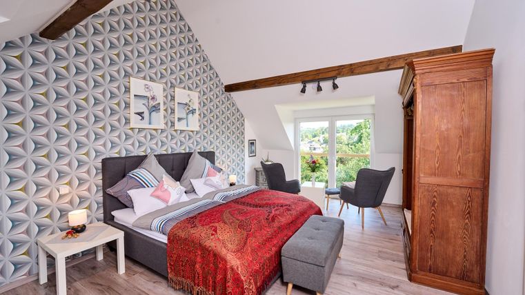 Ein modernes Schlafzimmer mit einem großen Bett und dekorativen Kissen. Die Wände sind mit einem geometrischen Muster versehen und es gibt einen Zugang zur Terrasse.
