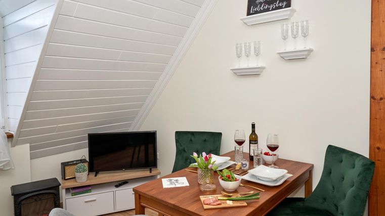 Ein gemütlicher Essbereich mit einem Tisch, zwei Stühlen und einer Fernsehecke. Auf dem Tisch stehen Wein und frische Blumen.