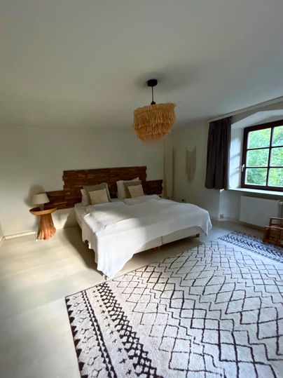 Ein helles Schlafzimmer mit einem großen Bett und natürlichem Holz. Ein gemütlicher Teppich und ein Fenster sorgen für eine einladende Atmosphäre.