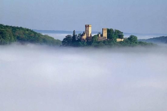 Eine beeindruckende Burg auf einem Hügel, umgeben von Nebel. Der Himmel ist klar und der Hintergrund zeigt sanfte Hügel.