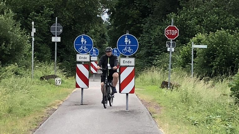 Ein Radfahrer fährt auf einem Weg, der durch Warnzeichen und Absperrungen markiert ist. Im Hintergrund sind Bäume und eine grüne Landschaft zu sehen.