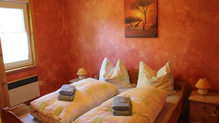 Ein gemütliches Schlafzimmer mit zwei Betten, die mit bunten Decken und Handtüchern dekoriert sind. Die Wände sind in warmen Orangetönen gestrichen und ein Bild mit einem Baum hängt über dem Bett.