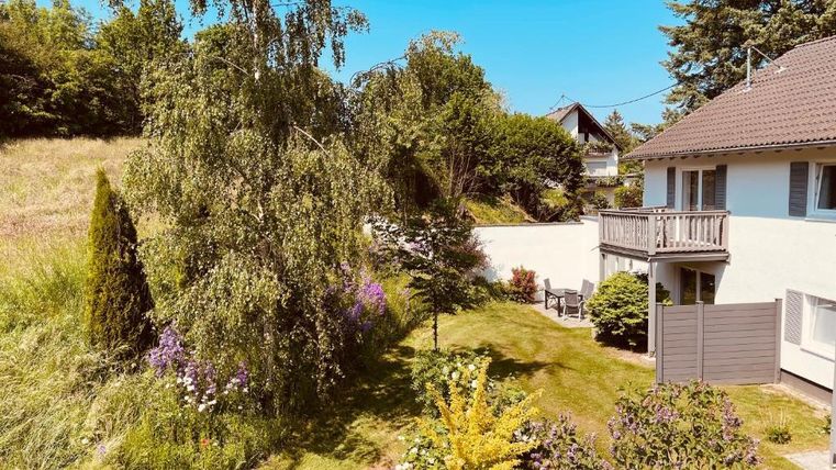 Ein schöner Garten mit unterschiedlichen Pflanzen und einem kleinen Teich. Im Hintergrund steht ein Haus mit einem Balkon.