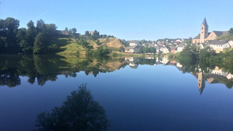 Ein ruhiger See spiegelt die Landschaft und das Dorf wider. Im Hintergrund sind sanfte Hügel und eine Kirche zu sehen.