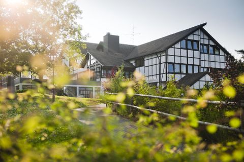 Ein charmantes Gebäude im Fachwerkstil, umgeben von Grün. Die Sonne scheint durch die Bäume und es gibt einen angenehmen, einladenden Eindruck.