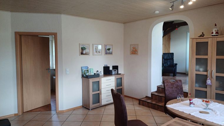 Eine helle und einladende Wohnung mit Fliesenboden. Im Raum befinden sich Möbel, Bilder an der Wand und eine Treppe, die zu einem weiteren Bereich führt.