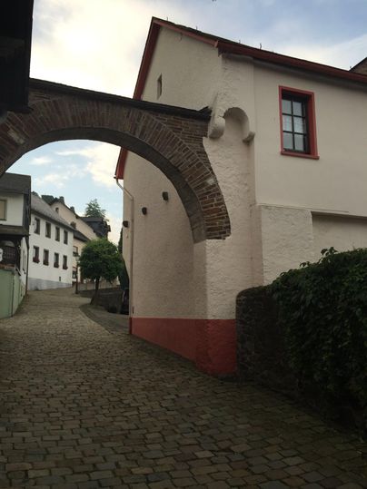 Eine enge Straße mit einem historischen Bogen und weißen Häusern. Die Umgebung ist ruhig und grün.