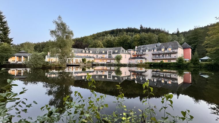 Ein ruhiges Hotelgebäude spiegelt sich im klaren Wasser eines Teiches. Im Hintergrund sind grüne Bäume und sanfte Hügel zu sehen.