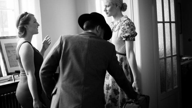 Drei Personen in einem hellen Raum, zwei Frauen unterhalten sich, während ein Mann ihnen zuhört. Eine der Frauen trägt ein Blumenmuster, die andere ein elegantes Outfit.