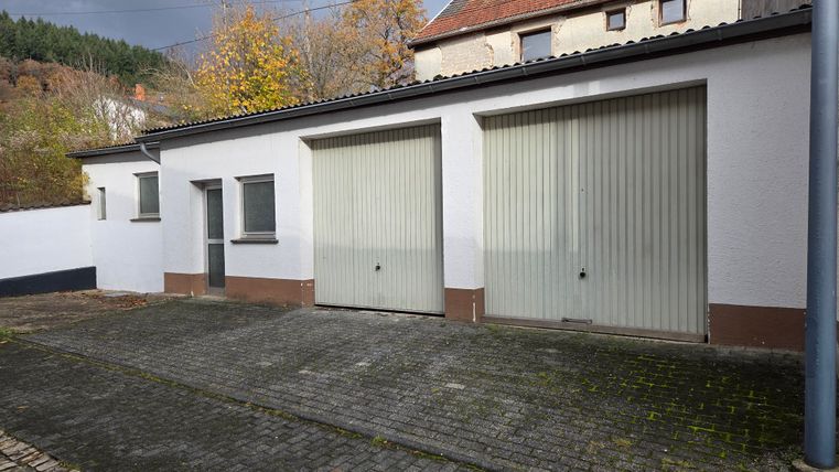 Ein Garagengebäude mit zwei großen, grauen Rolltoren. Im Hintergrund sind Bäume und ein weiteres Gebäude sichtbar.