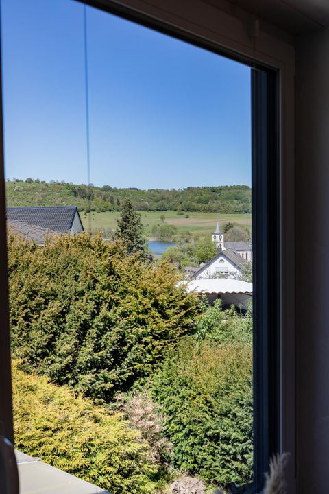 Blick aus einem Fenster auf eine grüne Landschaft mit Bäumen und einem kleinen See. Im Vordergrund sind Sträucher und Häuser sichtbar.