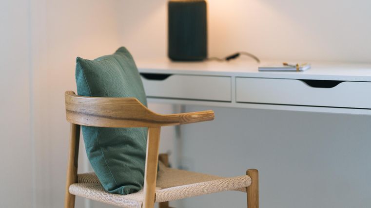 Ein moderner Schreibtisch mit einem Holzstuhl und einem Kissen. Im Hintergrund sind ein Leuchten und ein Lautsprecher sichtbar.